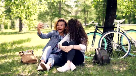 Cheerful-women-are-looking-at-smartphone-screen-and-laughing-then-taking-selfie-together-posing-for-camera-sitting-on-grass-in-park.-Bicycles-and-backpacks-are-visible.
