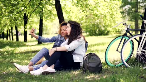 Mujer-joven-y-su-amiga-afroamericana-están-tomando-selfie,-posando-y-teniendo-diversión-sentados-en-el-césped-en-el-parque.-Día-asoleado-caliente,-hermosa-naturaleza-y-concepto-de-estilo-de-vida-moderno.