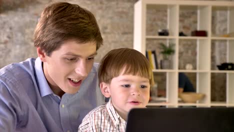 Young-father-holding-his-smiling-son-and-looking-at-laptop,-sitting-in-modern-office,-happy