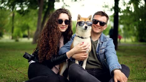 Portrait-of-cute-couple-and-shiba-inu-dog-all-wearing-sunglasses-looking-at-camera-and-smiling-sitting-on-grass-in-park.-Modern-lifestyle,-cute-animals-and-nature-concept.