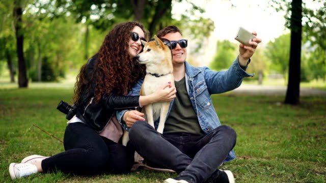 Handsome-young-man-is-taking-selfie-with-his-pretty-wife-and-cute-dog,-all-wearing-sunglasses.-Guy-is-holding-smartphone-taking-pictures-and-posing.