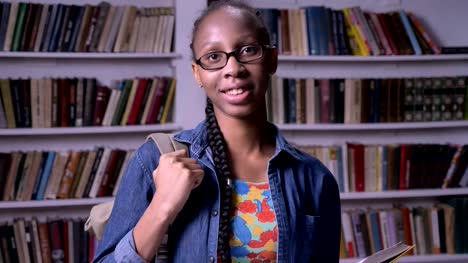 Young-beautiful-african-american-woman-in-glasses-reading-book-in-library-and-looking-at-camera,-happy,-smiling,-bookshelves-background