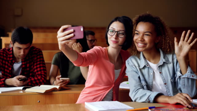 Fröhliche-Mädchen-Studenten-nehmen-Selfie-mit-Smartphone-sitzen-an-Tischen-am-College,-Frauen-posieren-Hand-Gesten-und-umarmt.-Freunde-und-Technologiekonzept.