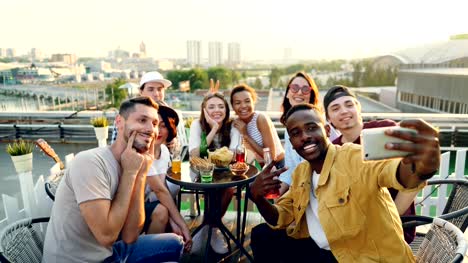 Hombre-joven-afroamericano-es-celebración-de-smartphone-y-selfie-con-su-grupo-de-amigos-y-multiétnica-con-botellas-y-copas-disfrutando-de-fiesta-en-la-azotea.