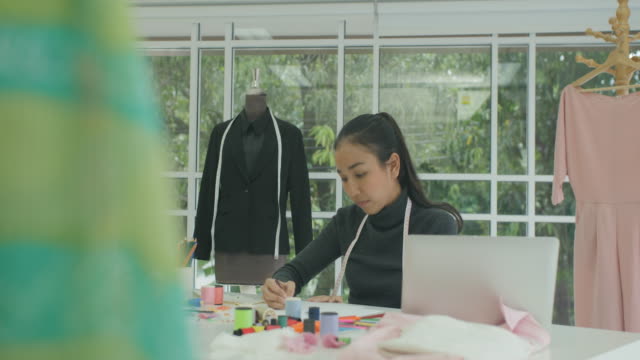DS-MS-schöne-Asiatin-Modedesignerin-sitzt-an-ihrem-Schreibtisch-auf-einem-Laptop-in-modernes-Studio-mit-Blick-auf-Skizzen,-Notizen