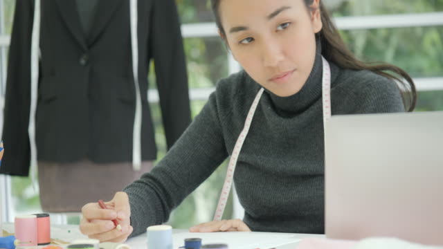 Hermosa-mujer-asiática-moda-diseñador-sentado-en-su-escritorio-mirando-bocetos-en-un-ordenador-portátil-en-estudio,-tomando-notas
