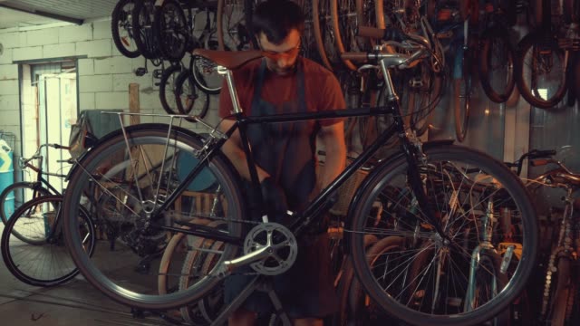 Thema-Kleinunternehmen-Fahrradreparatur.-Ein-junger-kaukasischen-Brünette-Mann-mit-Sicherheit-Schutzbrillen,-Handschuhe-und-eine-Schürze-nutzt-ein-Handwerkzeug-zu-reparieren-und-stellen-das-Fahrrad-in-der-Garage-Werkstatt