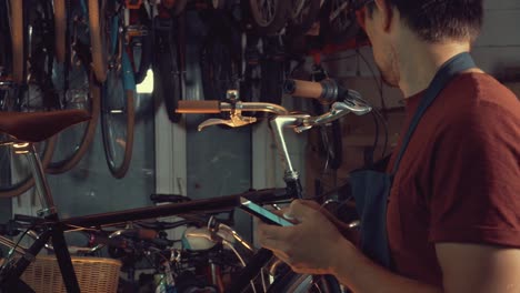 Thema-Kleinunternehmen-Fahrradreparatur.-Kaukasische-Brünette-Jüngling-tragen,-Schutzbrille,-Handschuhe-und-Fartukhe-Handy-Technologie-nutzt,-macht-sich-Notizen,-Checkliste-in-Fahrradwerkstatt