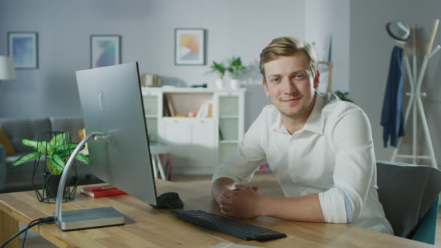 Portrait-of-the-Handsome-Young-Man-Looking-at-the-Camera-While-Sitting-at-His-Desk-with-Personal-Computer.-In-the-Background-Cozy-Living-Room.