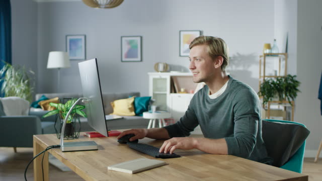 Portrait-of-the-Focused-Young-Man-Working-on-a-Personal-Computer-while-Sitting-at-His-Desk.-In-the-Background-Cozy-Living-Room.