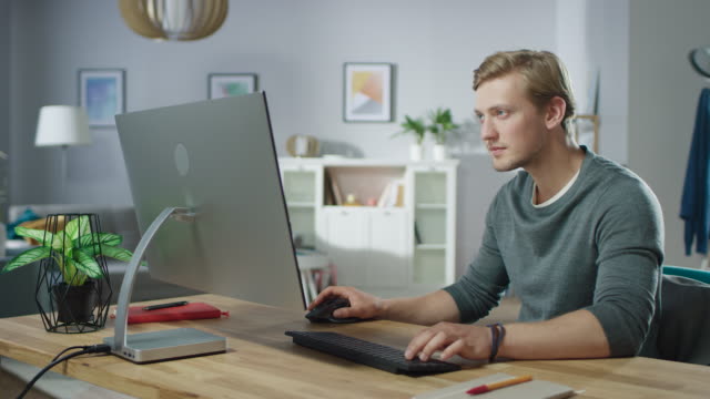 Portrait-of-the-Handsome-Man-Working-on-Personal-Computer-while-Sitting-at-His-Desk.-In-the-Background-Stylish-Cozy-Living-Room.-Young-Freelancer-Working-From-Home.