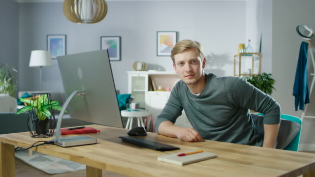 Portrait-of-the-Handsome-Young-Man-Looking-at-the-Camera-While-Sitting-at-His-Desk-with-Personal-Computer.-In-the-Background-Cozy-Living-Room.