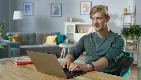 Gut-aussehend-lächelnde-jungen-Mann-mit-Laptop-beim-Sitzen-am-Schreibtisch-von-seiner-gemütlichen-Wohnzimmer.