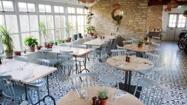 Runde-Tische-und-Entenei-blaue-Stühle-in-einem-leeren-Restaurant-mit-gemusterten-Bodenfliesen-und-Zimmerpflanzen-auf-der-Fensterbank-wachsen