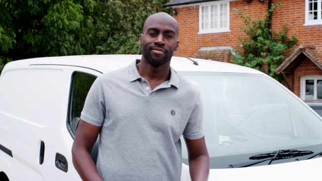 Young-adult-black-tradesman-standing-and-leaning-on-his-white-van-smiling-to-camera-crosses-his-arms,-close-up
