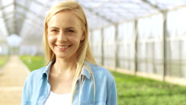 On-a-Sunny-Day-Beautiful-Blonde-Gardener-Stands-Smiling-in-a-Greenhouse-Full-of-Colorful-Flowers.