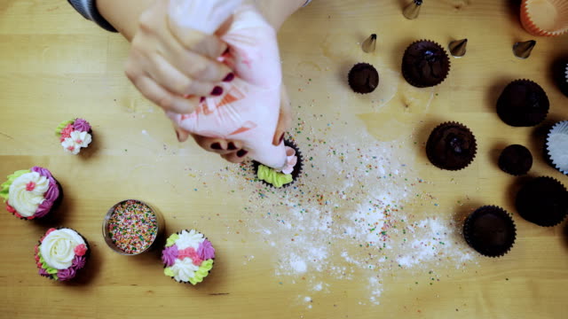 Vista-superior-de-la-mujer-manos-decorando-los-cupcakes-de-chocolate.-Mujer-joven-con-la-Manga-pastelera-para-color-crema