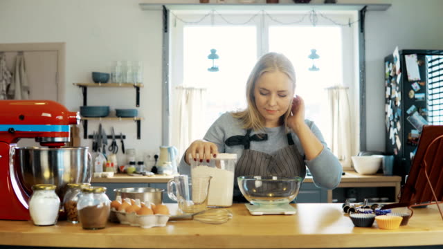 Young-blonde-woman-using-the-kitchen-scales-to-weighing-the-flour.-Beautiful-female-baking-the-cupcakes-on-the-kitchen