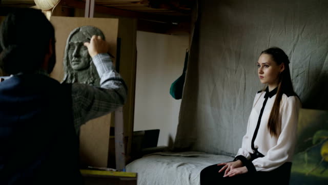 Bildhauer-Skulptur-des-menschlichen-Gesichts-auf-Leinwand-während-die-junge-Frau-posiert-mit-ihm-im-Kunstatelier-erstellen