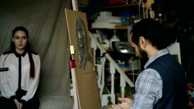 Bildhauer-Skulptur-des-menschlichen-Gesichts-auf-Leinwand-während-die-junge-Frau-posiert-mit-ihm-im-Kunstatelier-erstellen