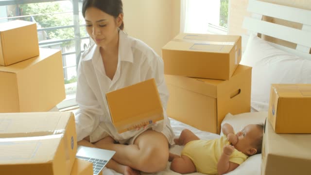 Joven-madre-asiática-y-su-niño-recién-nacido,-comprobación-orden-del-ordenador-portátil-para-el-cliente-y-entrega-en-línea-de-embalaje-listo-en-dormitorio.