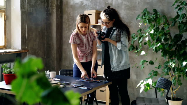 Young-female-enterpreneurs-are-placing-photos-on-table-to-make-flat-lay-and-shooting-with-camera-while-standing-in-modern-office.-Women-are-sharing-ideas-and-discussing-design.