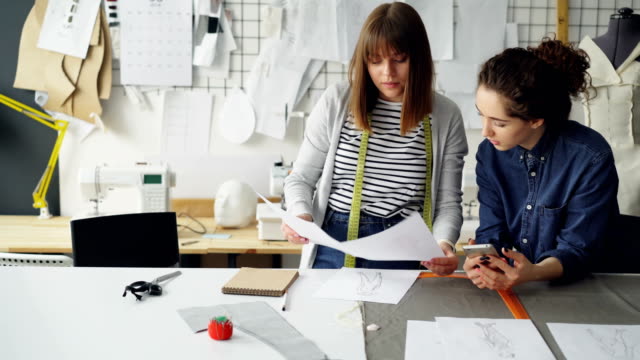 Junge-Frauen-in-Industrie-Design-sind-Skizzen-zu-vergleichen-und-zu-reden,-während-der-Arbeit-in-modernen-Schneiderei.-Produktive-Teamarbeit-Konzept.