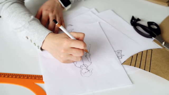 Close-up-shot-of-woman's-hand-drawing-outlines-of-stylish-ladies'-garment.-Paper,-tailor's-scissors,-ruler-and-coffee-are-visible-on-studio-table.-Designer-at-work-concept.