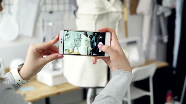 Close-up-shot-of-women's-hands-holding-smartphone-and-photographing-tailoring-dummy-with-half-finished-garment-pinned-to-it.-Modern-technologies-in-small-business-concept.