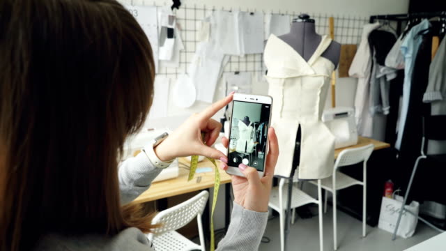 Young-female-clothing-design-blogger-is-shooting-tailoring-dummy-with-half-finished-garment-pinned-to-it.-Close-up-shot-of-girl's-hands-and-smartphone.
