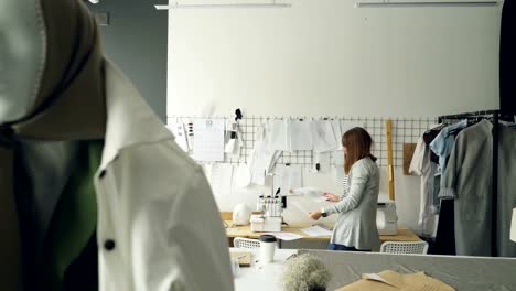 Attractive-woman-clothing-designer-is-looking-at-garment-sketches-hanging-on-wall-then-choosing-new-drawings.-Mannequin-dresses-in-trendy-clothes-is-in-foreground.