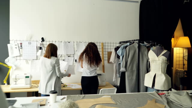 Young-entrepreneurs-clothing-designers-standing-in-front-of-wall-with-hanging-sketches,-discussing-drawings-and-placing-images-with-clips.-Creative-team-working-together-concept.