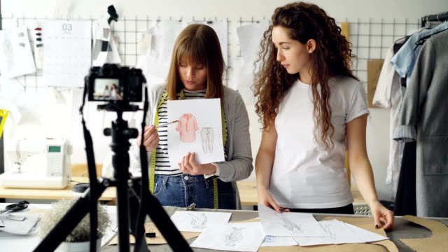 Young-women-fashion-bloggers-recording-video-blog-about-ladies'clothes-on-camera-and-talking-to-followers-in-modern-studio.-Many-garment-sketches-are-visible.