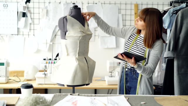 Diseño-moda-joven-empresario-está-trabajando-con-la-tableta-mientras-revisa-prendas-en-maniquí-de-sastrería.-Tecnología-moderna-en-el-concepto-de-fabricación-de-ropa.