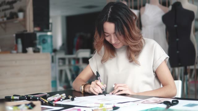young-fashion-designer-girl-makes-a-sketch-in-pencil-at-the-atelier.-business-woman-with-Asian-appearance-working-in-the-office