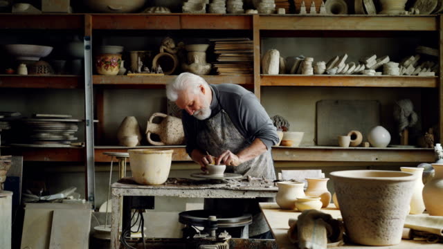 Erfahrener-Keramiker-schafft-Geschirr-aus-Ton-auf-der-Töpferscheibe-in-Werkstatt.-Herstellung-von-Eathenware,-handgemachten-Utensilien-und-professionelle-Keramik-Konzept.