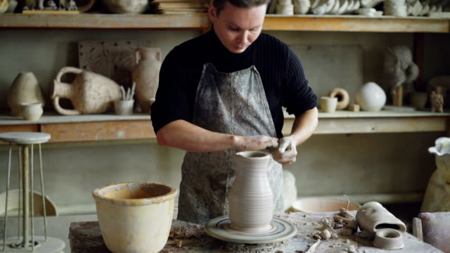 Handsome-young-man-is-molding-ceramic-vase-from-clay-on-spinning-throwing-wheel-while-working-in-potter's-studio.-Professional-is-concentrated-on-work.
