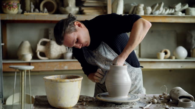 Skillful-young-potter-is-molding-ceramic-vase-from-clay-on-turning-throwing-wheel-while-working-in-potter's-workshop.-Professional-is-concentrated-on-work.