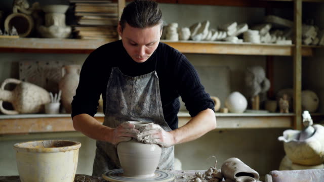 Experienced-ceramist-is-forming-ceramic-vase-from-clay-on-spinning-throwing-wheel-while-working-in-potter's-workshop.-Professional-is-focused-on-work.