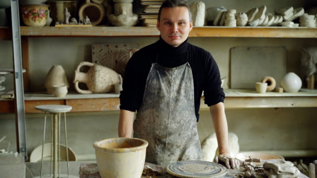 Portrait-of-handsome-young-potter-in-muddy-apron-standing-at-table-in-workshop-and-looking-at-camera.-Shelves-with-handmade-vases-and-pots-in-background.