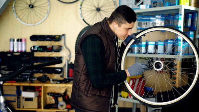 Young-serviceman-is-cleaning-bicycle-wheel-outside-and-inside-with-tools-and-piece-of-cloth-and-listening-to-music-with-earphones.-Profession-and-people-concept.