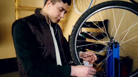 Mecánico-autónomo-joven-es-fijación-de-rueda-de-bicicleta-en-el-taller.-Joven-lleva-ropa-casual,-chaleco-cálido-y-está-escuchando-música-con-auriculares.