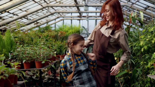 Young-female-gardener-in-apron-and-her-cute-daughter-are-dancing-in-greenhouse-having-fun.-Happy-family,-gardening,-parents-and-children-concept.