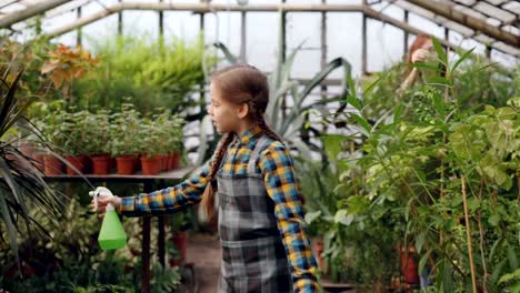 Pretty-girl-is-walking-in-greenhouse-and-spraying-water-on-greenery-while-her-busy-mother-is-working-in-background.-Childhood,-helping-parents-and-family-concept.