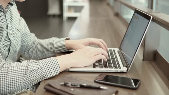 Hombre-de-negocios-asiático-joven-usando-la-computadora-portátil-en-el-espacio-de-trabajo-con-el-smartphone-y-el-portátil-en-el-escritorio-de-madera.-Macho-mano-escribiendo-en-el-teclado-del-ordenador-portátil.-Estilo-de-vida-independiente-en-concepto-