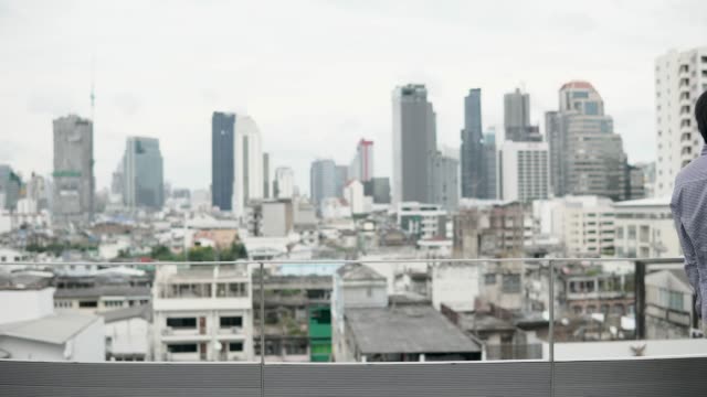 Young-Asian-businessman-using-smartphone-for-business-talk-walking-on-office-building-rooftop-terrace-with-city-view-in-the-background.-Business-communication-and-corporation-concepts.