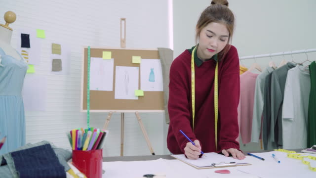 Junge-asiatische-Frau-Mode-Designer-Zeichnung-mit-Bleistift-und-Papier-während-der-Arbeit-im-Workshop-Studio-betrachten.