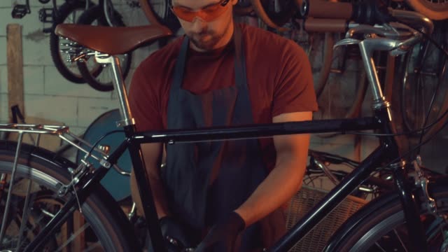 theme-small-business-bike-repair.-A-young-Caucasian-brunette-man-wearing-safety-goggles,-gloves-and-an-apron-uses-a-hand-tool-to-repair-and-adjust-the-bike-in-the-workshop-garage
