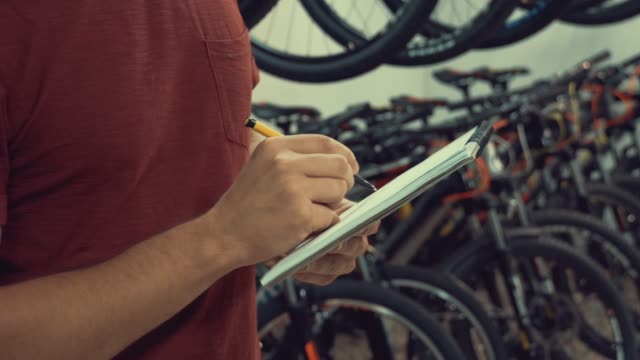 Thema-der-Kleinunternehmen,-die-Fahrräder-zu-verkaufen.-Junge-kaukasischen-männlichen-Brünette-Kleinunternehmer,-Filialleiter-verwendet-Notizblock-und-Stift-macht-Noten,-Checkliste-im-Fahrradladen