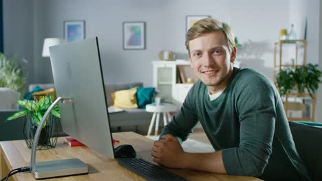 Retrato-de-la-centra-joven-hombre-trabajando-en-un-ordenador-Personal-mientras-que-sentado-en-su-escritorio,-sonriente-y-mirando-a-la-cámara.-En-el-fondo-acogedora-sala-de-estar.